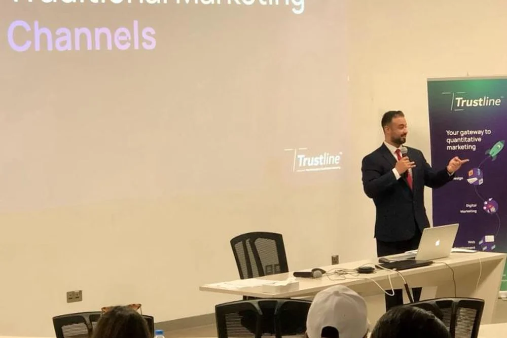Ali Owji presenting to a seated audience beside a Trustline banner, a slide behind him reading “Traditional Marketing Channels”.