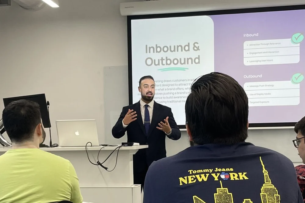 Ali Owji lecturing to students, a slide behind him comparing inbound and outbound marketing.